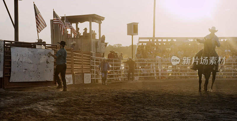在一个骑牛/牛仔竞技活动中，一个骑着马的牛仔穿过一个挤满了人的体育场，另一个牛仔在日落时把一个覆盖物戴在了公牛的围栏上